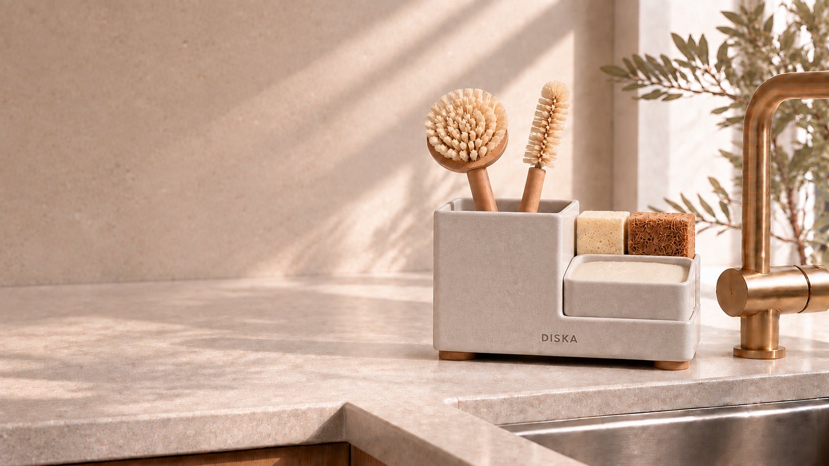 The Diska Stand in Stone on a limestone worktop by the sink, holding two brushes, two sponges and dish paste